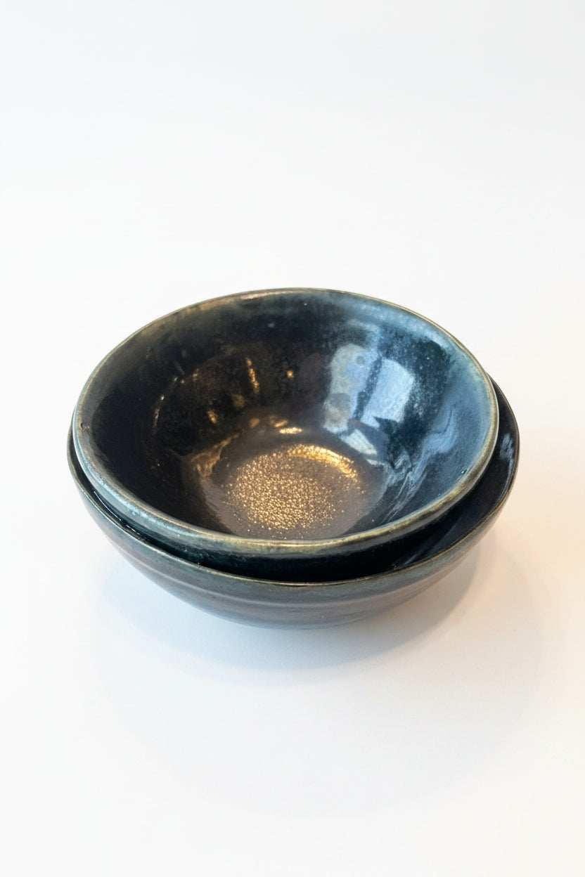 A small handmade ceramic bowl with a glossy black glaze and a visible gold-toned center reflection, resting on a scratched dark metallic surface.