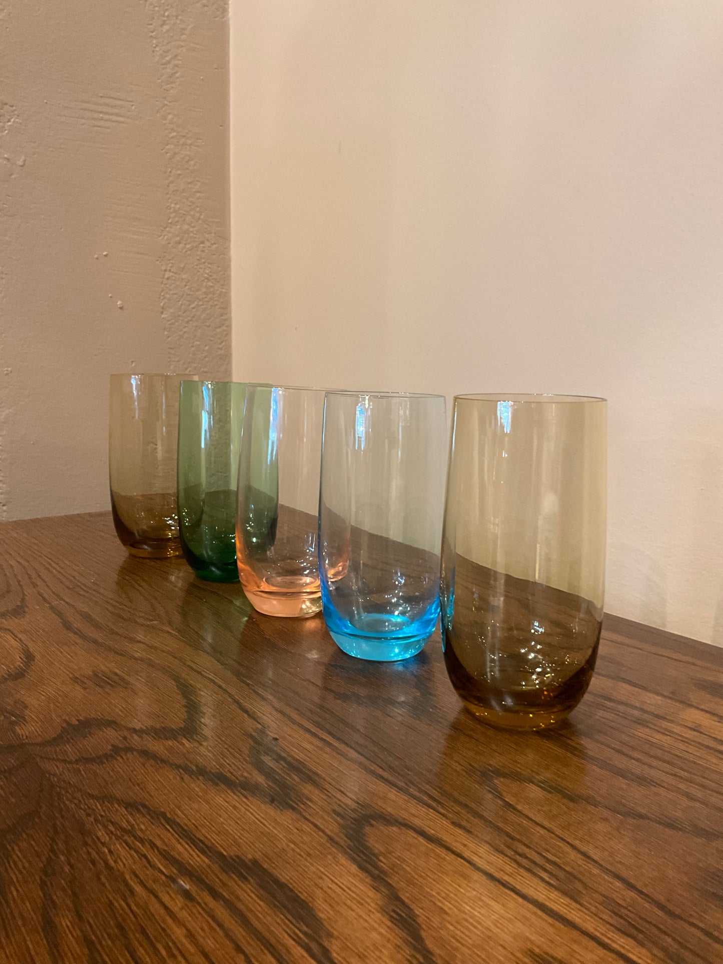 Set of five tall drinking glasses in various colors from The Vault Creative, shown filled with clear liquid and set on a table.