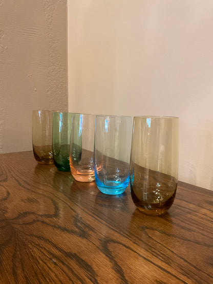Set of five tall drinking glasses in various colors from The Vault Creative, shown filled with clear liquid and set on a table.